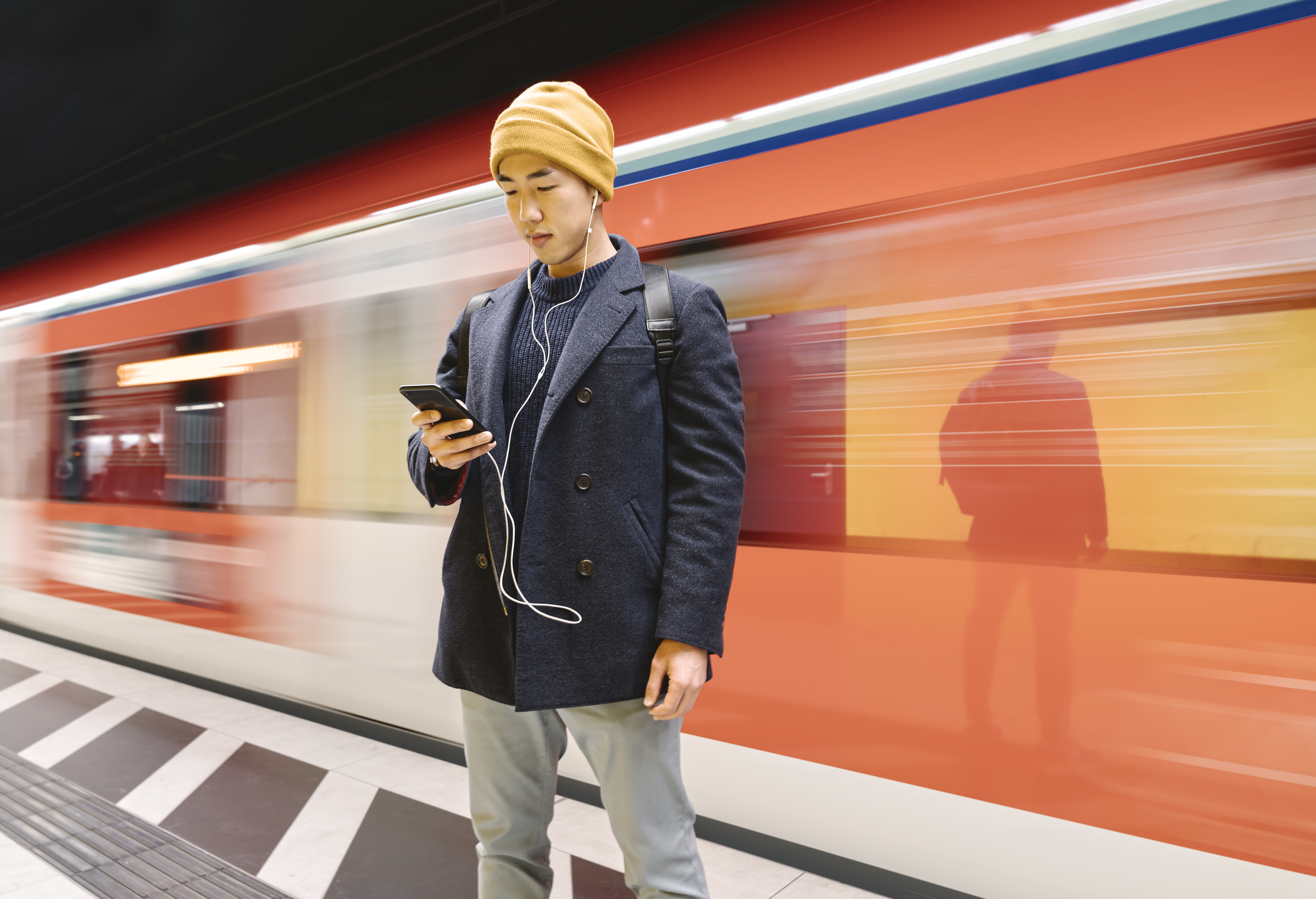 Asian man wearing a cap looking at his smartphone in front of a moving train.